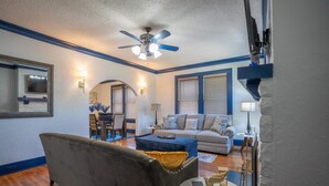 Un salon avec un canapé gris, un pouf bleu, une petite table ronde et un ventilateur de plafond.