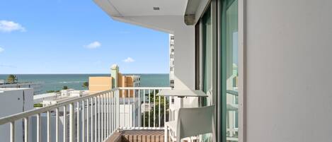 A balcony with a view of the ocean, a white railing, and a table with a chair.