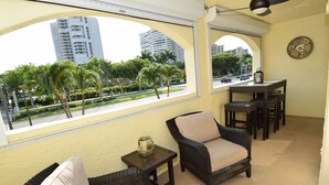 Een balkon met rieten meubels, een tafeltje en uitzicht op palmbomen en gebouwen.