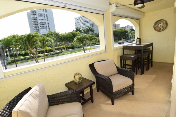 Un balcón con mobiliario de mimbre, una pequeña mesa y vistas a palmeras y edificios.