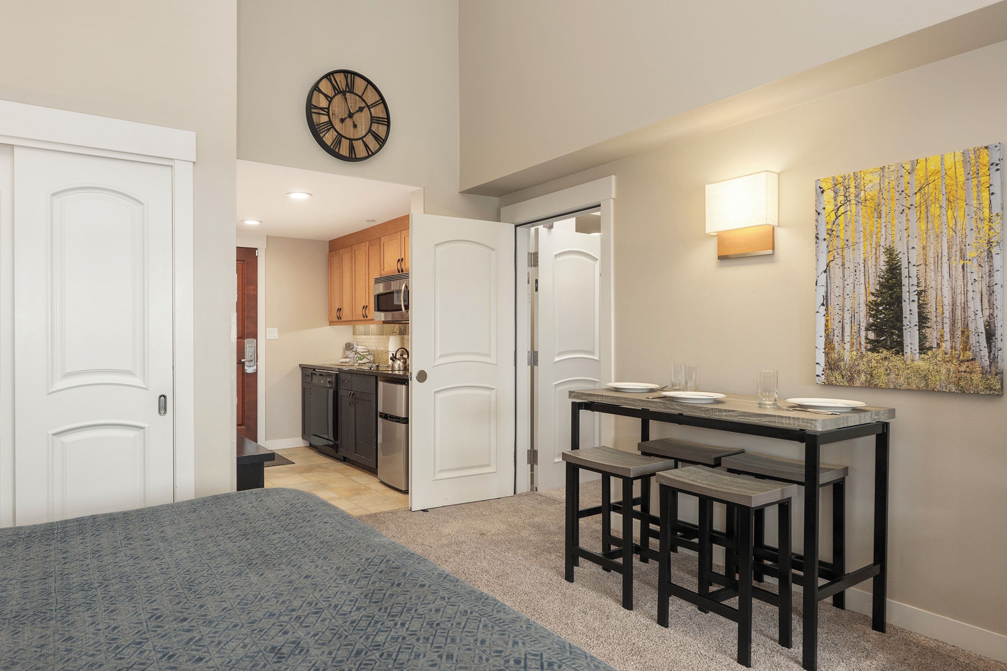 A bedroom with a bed, a dining area with a table and chairs, a kitchen with cabinets and appliances, and a wall clock.