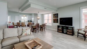 A spacious living room with a large sofa, a coffee table, and a TV on a stand. There is a dining area with a table and chairs, and a kitchen in the background. - 🌞Oceanfront Rooftop getaway 🏖️Steps to sand 🐶Dog Friendly⚡️EV charger (North Topsail Beach)
