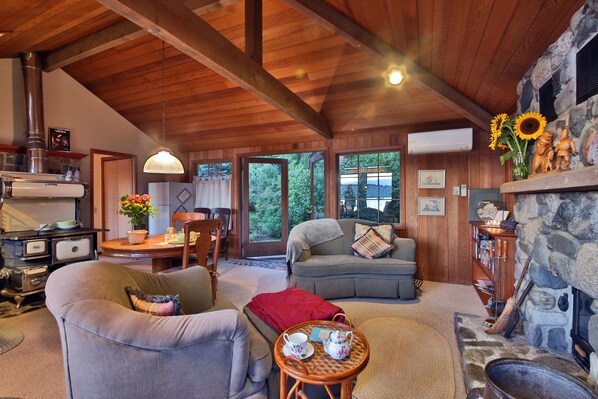 A cozy living room with a fireplace, wooden ceiling beams, a dining area with a table and chairs, and a view of the outdoors through large windows.