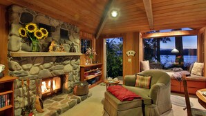 A cozy cabin interior with a stone fireplace, wooden shelves, a comfortable seating area, and a view of the outdoors.