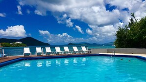 Una piscina con sillones blancos, un cielo azul despejado y vistas al mar y montañas al fondo.