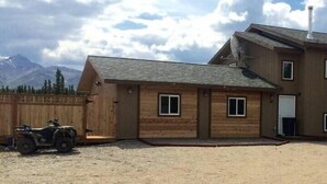 A two-story wooden building with a garage, a covered driveway, and a mountain backdrop. - Denali Park 2 Bedroom Mom in Law (Healy)