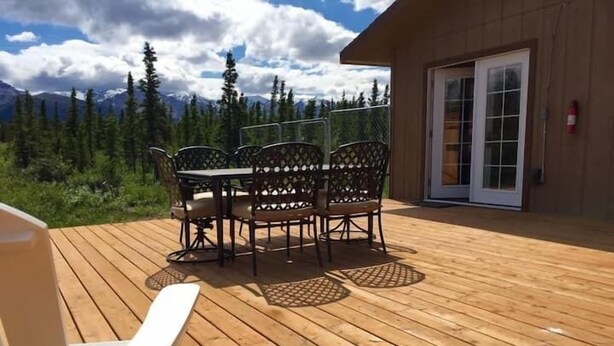 A wooden deck with a table and chairs, a white chair, and a building with a glass door. - Denali Park 2 Bedroom Mom in Law (Healy)