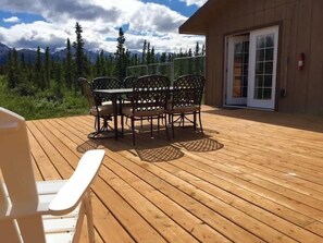 A wooden deck with a table and chairs, a white chair, and a building with a glass door. - Denali Park 2 Bedroom Mom in Law (Healy)