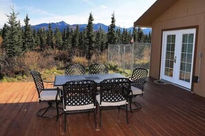 An outdoor patio with a table and chairs, surrounded by a wooden deck and a view of mountains and trees.