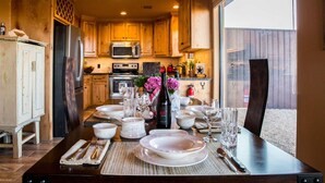 A dining table set with plates, glasses, and cutlery, a bottle, and a vase with flowers in a kitchen with wooden cabinets and a large window. - Comanche cabin on the west rim (Canyon)