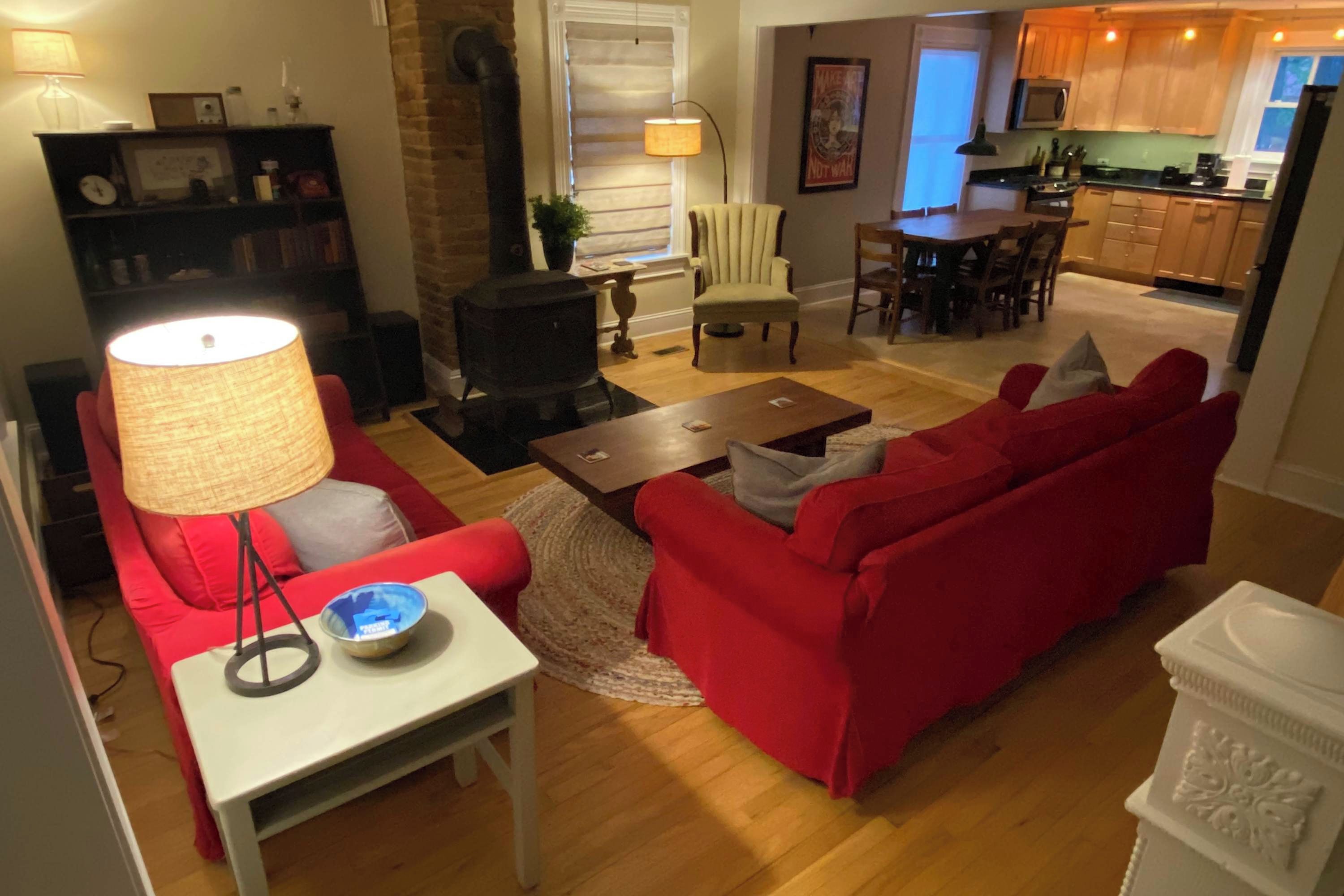 A living room with a red sofa, a white table, and a lamp. There is a kitchen in the background with wooden cabinets and a dining area.