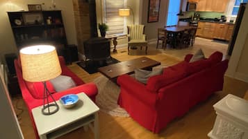 A living room with a red sofa, a white table, and a lamp. There is a kitchen in the background with wooden cabinets and a dining area.