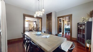 Un coin repas avec une table en bois, des chaises et un lustre. On aperçoit une cuisine au fond.