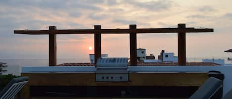 Una terraza en la azotea con una parrilla, sillones de descanso y vistas a la puesta de sol sobre el mar.