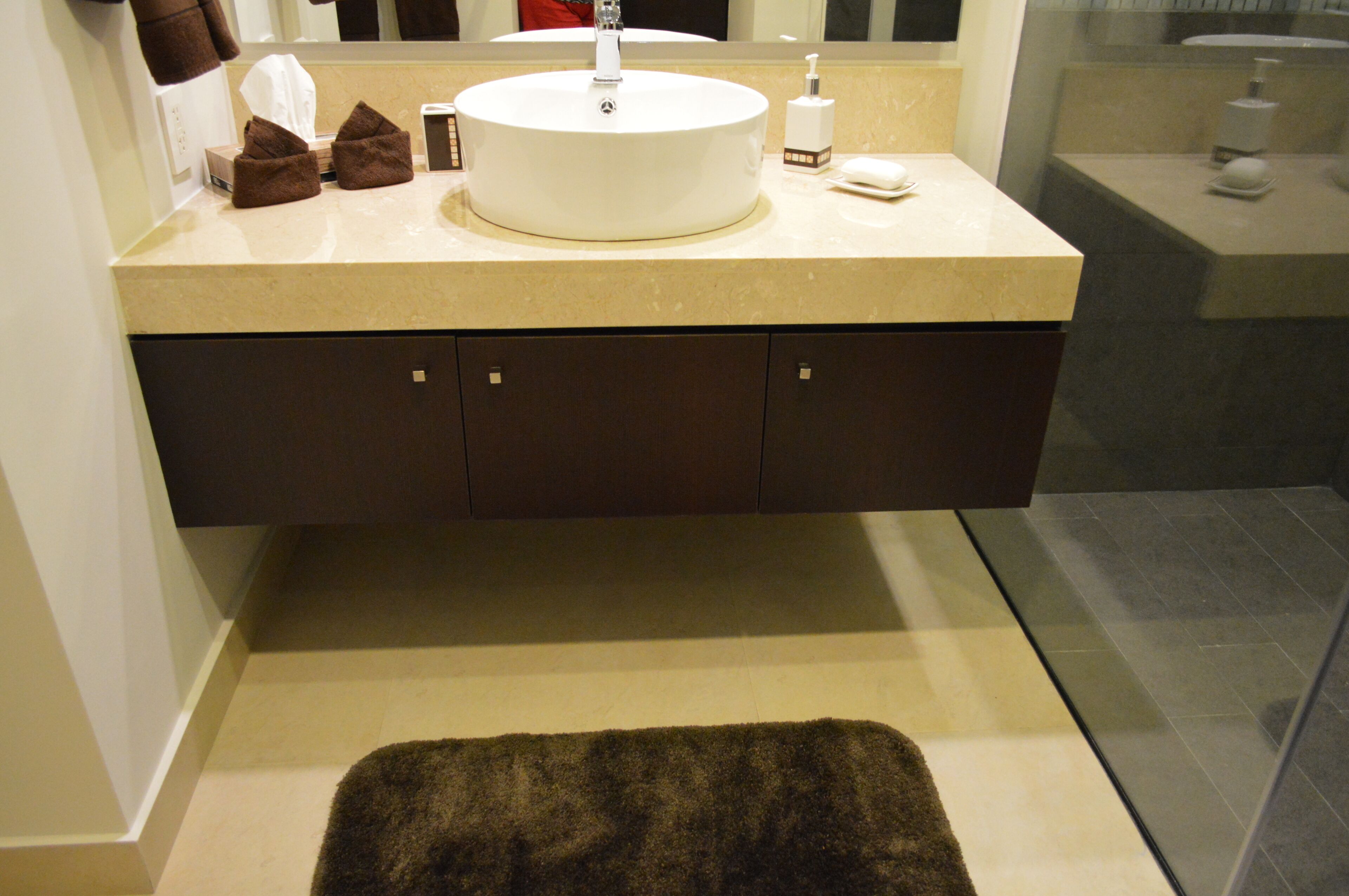 Une salle de bain avec un lavabo rond, des meubles sombres, un plan de toilette beige et un tapis marron sur le sol.