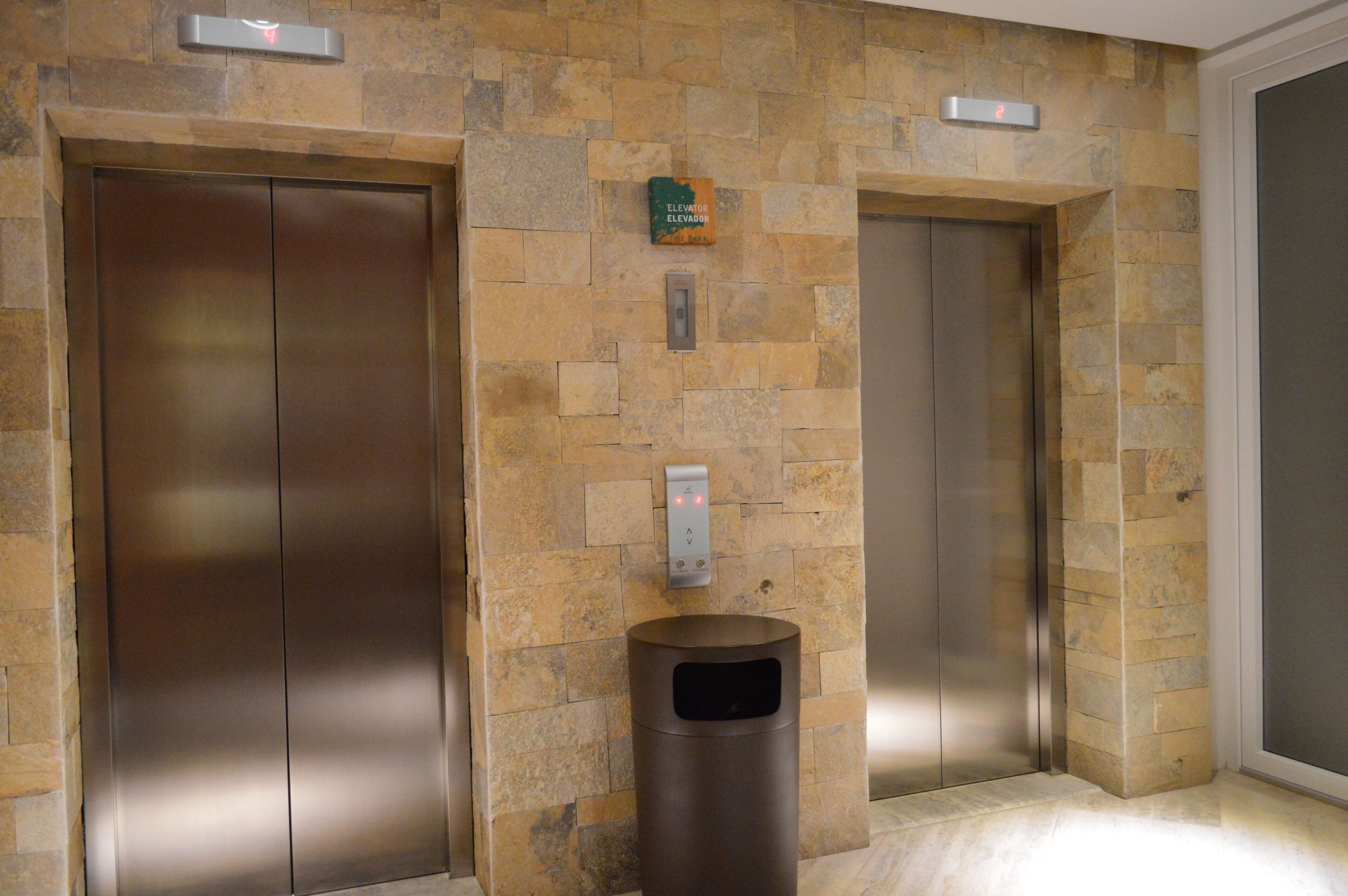 Deux portes d’ascenseur, un mur en pierre et une poubelle dans le hall d’entrée.