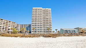 A beachfront hotel with multiple balconies, a sandy beach in the foreground, and a clear blue sky.
