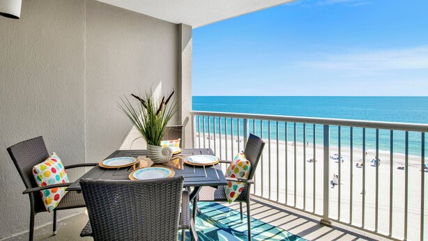 A balcony with a view of the beach, featuring a table with plates and chairs, a potted plant, and a railing.