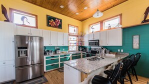 A modern kitchen with a stainless steel refrigerator, white cabinets, teal accents, granite countertops, and a central island with bar stools. - Serenity Sands Beach HouseFamily-Friendly Ocean View Home (PORT ARANSAS)
