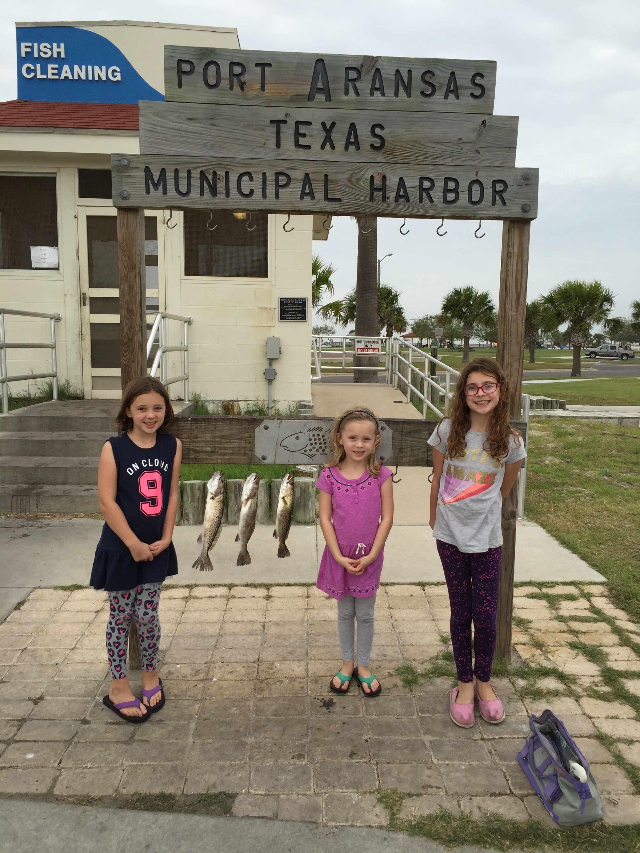 Three individuals standing in front of a wooden sign that reads 'Port Aransas Texas Municipal Harbor.'