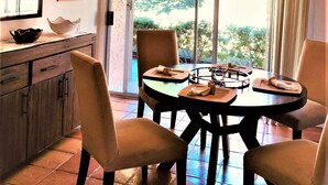 A dining area with a round table, four chairs, a hanging light fixture, a window with a view of greenery, and a wooden cabinet with a bowl and a framed picture.