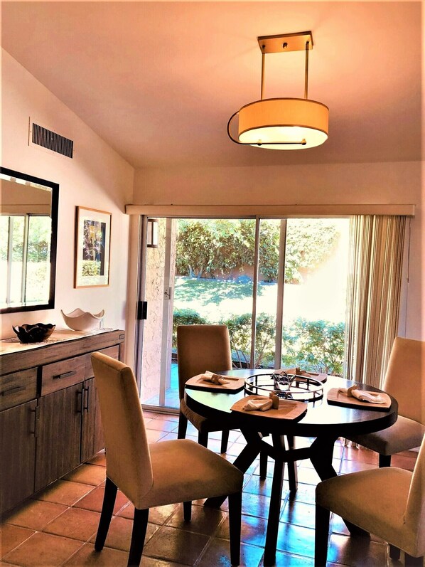 A dining area with a round table, four chairs, a hanging light fixture, a window with a view of greenery, and a wooden cabinet with a bowl and a framed picture. - Zen Pool Villa - Original Art & Vintage Design - Pool & Jacuzzi - Ironwood (Palm Desert)
