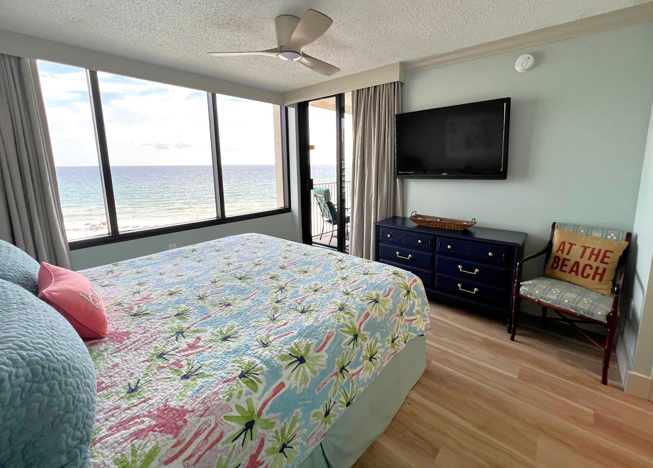 A bedroom with a large bed, a TV mounted on the wall, a chair with a beach-themed pillow, and a view of the ocean through the window.