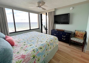 Ein Schlafzimmer mit einem großen Bett, einem an der Wand montierten Fernseher, einem Sessel mit einem Strandkissen und Blick auf den Ozean durch das Fenster.