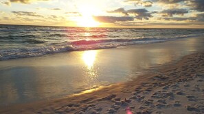 Une plage où l’on voit des empreintes de pas menant à l’eau, des vagues et un coucher de soleil.