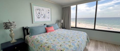 Un dormitorio con cama, ventilador de techo, mesita de noche, lámpara, un cuadro enmarco y un gran ventanal con vista a la playa.