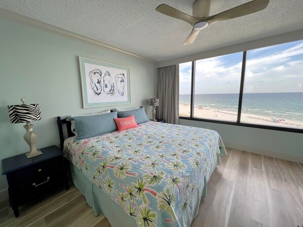 Une chambre avec un lit, un ventilateur de plafond, une table de chevet, une lampe, un tableau encadré et une grande fenêtre donnant sur la plage.