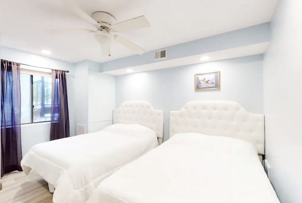 Quarto de hotel com duas camas, um ventilador de teto, uma janela com cortinas e um quadro na parede.