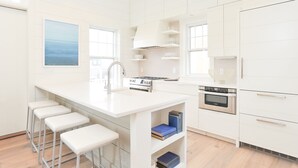 A modern kitchen with white cabinetry, a central island with bar stools, a stove, oven, and a large window providing natural light. - Luxurious Harbor “Elizabeth” Cottage! Walk & bike to everything (Nantucket)
