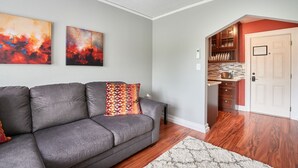 A living room with a gray sofa, two abstract paintings, a white door leading to a kitchen with wooden cabinets and a microwave.