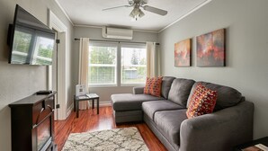 A living room with a gray sectional sofa, two patterned throw pillows, a flat-screen TV, a ceiling fan, and a large window with blinds.