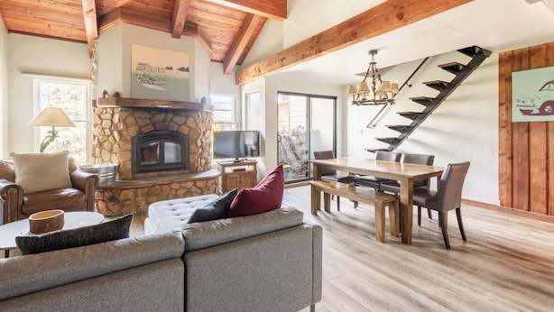 A spacious living room with a stone fireplace, wooden beams, a dining area, and a staircase.