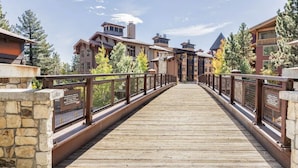 A wooden bridge with stone walls and metal railings, leading to a building with multiple stories and a sloped roof.