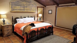 A bedroom with a wooden bed, two bedside tables with lamps, a large window with blinds, and a suitcase on the floor.
