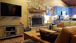 A living room with a stone fireplace, a television, a kitchen with blue cabinets, and a sofa.