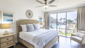 Un espacioso dormitorio con una cama grande, ventilador de techo y vistas a una piscina y palmeras.