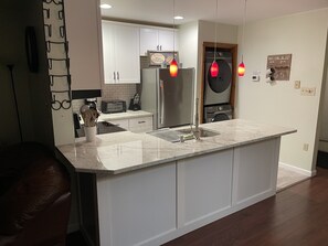 Una cocina moderna con armarios blancos, un refrigerador de acero inoxidable, horno empotrado y un lavamanos. Hay lámparas colgantes y una cafetera sobre la encimera.