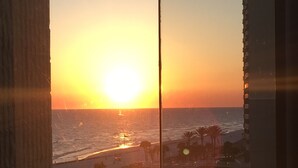 A sunset view from a window overlooking a beach with palm trees and buildings in the background. - Beautiful Beachfront Studio with Beach Chairs for 2 (Panama City Beach)