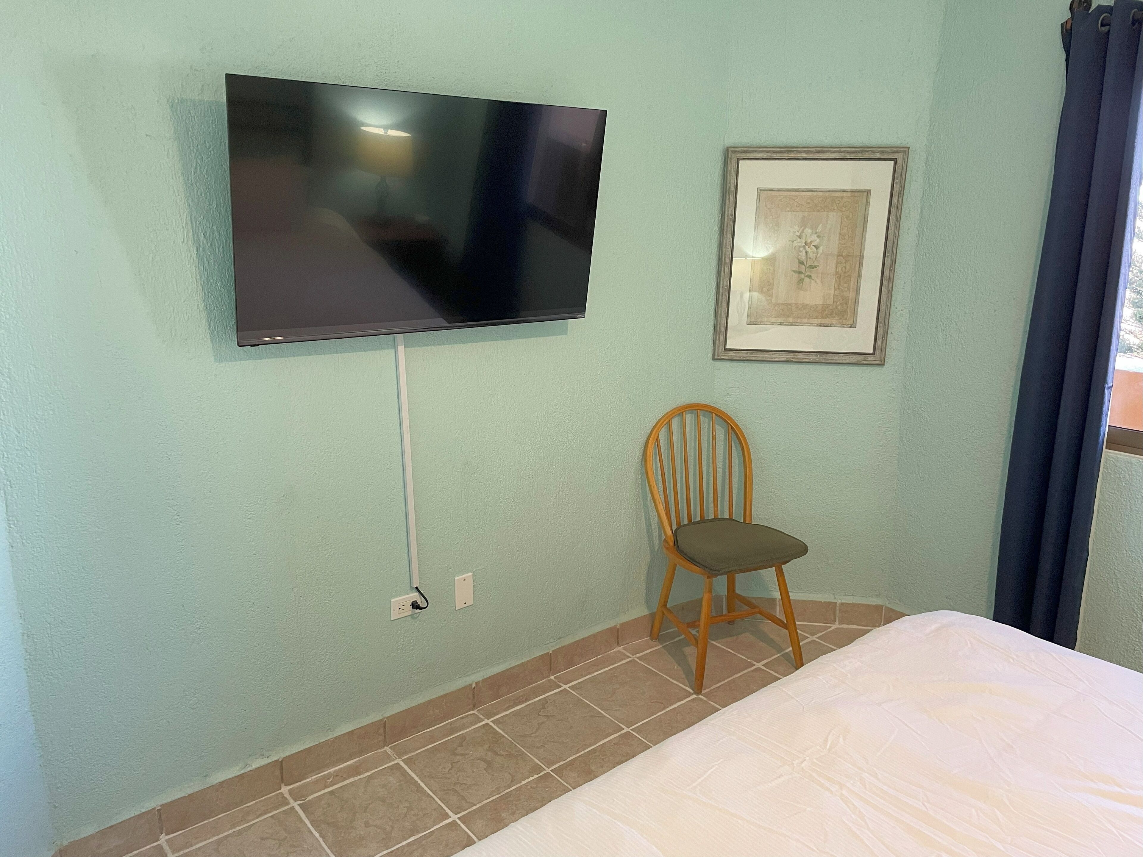A room with a bed, a television mounted on the wall, a chair, and a framed picture.