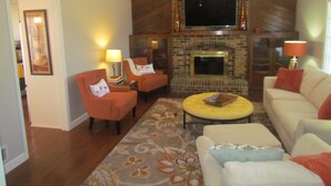 A living room with a fireplace, two orange armchairs, a yellow ottoman, and a white sofa.