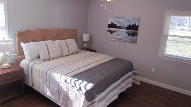 A bedroom with a bed, a nightstand, a lamp, a ceiling fan, a window, and a wall-mounted picture.