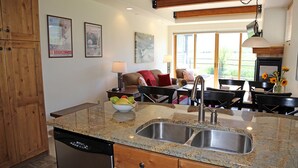 Una cocina con encimeras de granito, pileta de acero inoxidable y muebles de madera. Hay un comedor con mesa y sillas, y una ventana que proporciona luz natural.