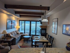 Un espacio de sala acogedor con sofá, mesa de comedor y sillas, chimenea y un ventanal con vistas a un jardín.