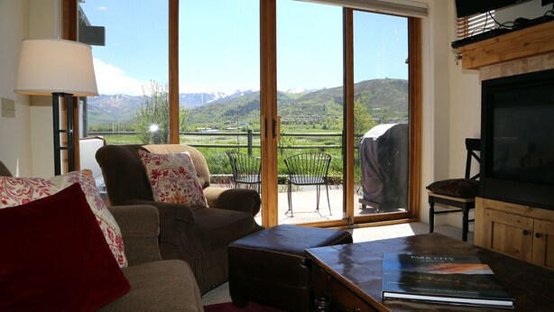 Una sala de estar con un sofá, una mesa de centro y un televisor. Se observa un ventanal con vistas a montañas y un jardín.