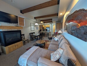 A modern living room with a fireplace, a sofa, a dining table, and a kitchen in the background.