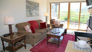 A living room with a sofa, armchairs, a coffee table, and a TV. There is a window with a view of greenery outside.